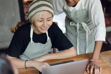 From above of optimistic diverse female workers of cafeteria discussing business plan while using laptop and taking notes in piece of paper at table