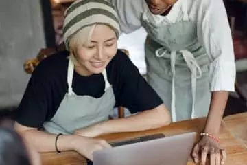 From above of optimistic diverse female workers of cafeteria discussing business plan while using laptop and taking notes in piece of paper at table