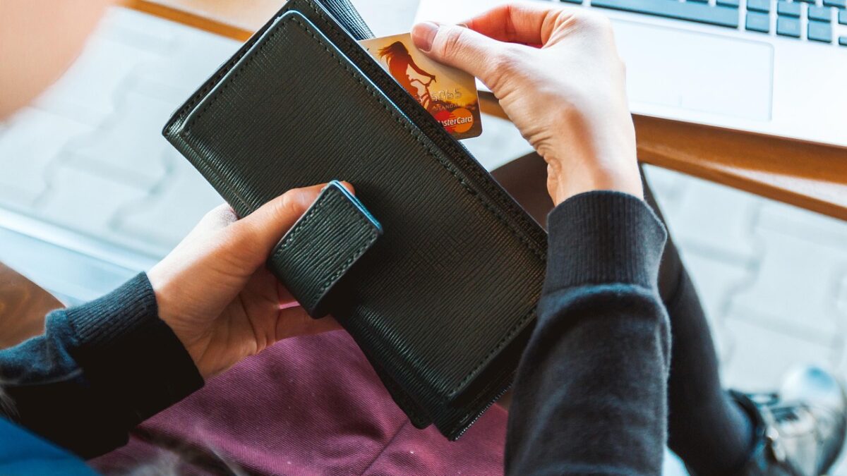 Une personne glisse une carte bancaire Mastercard dans un portefeuille noir ; un ordinateur portable est posé sur une table en arrière-plan.