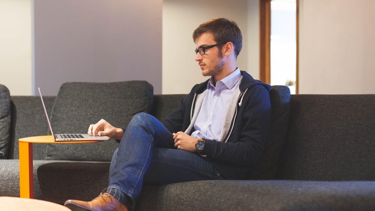 Un homme à lunettes, en veste bleu marine et jean, est assis sur un canapé gris et travaille sur un ordinateur portable posé sur une petite table ronde orange. Une table basse rose est au premier plan.