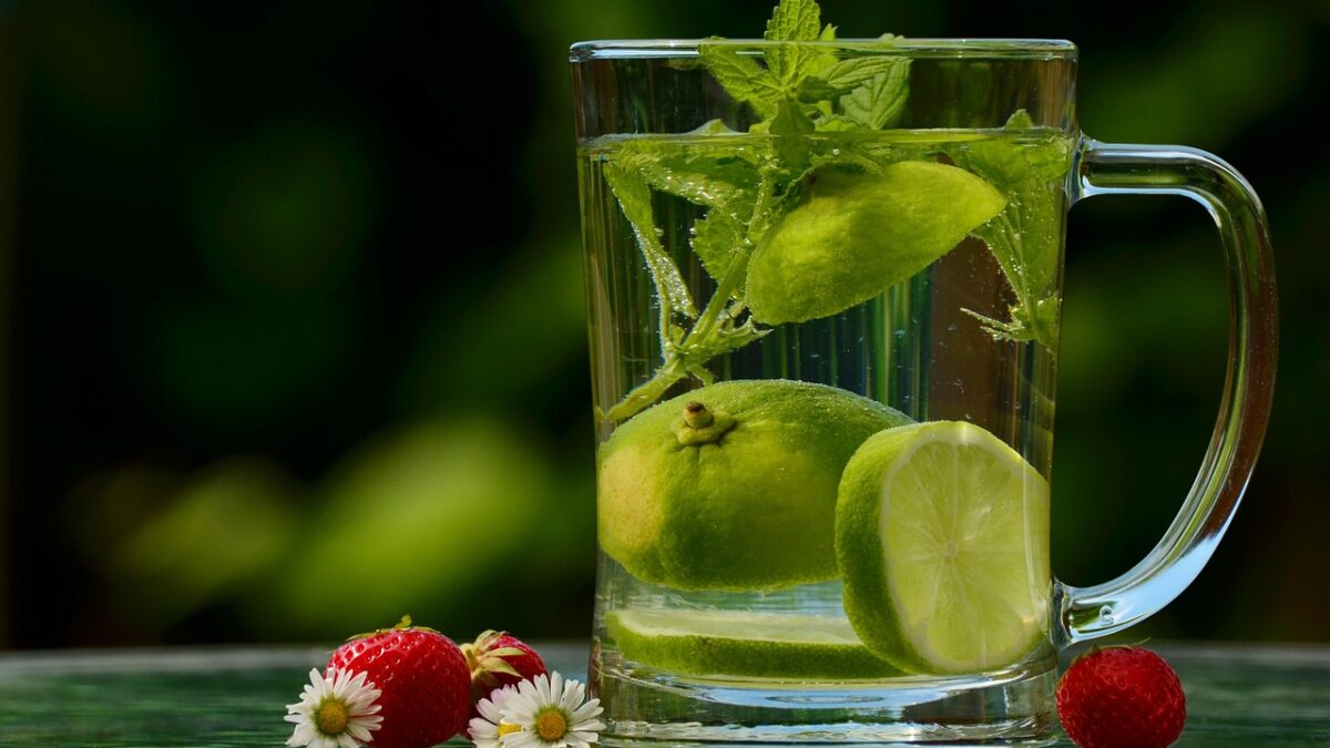 Un verre à anse rempli d'eau avec des tranches et un quartier de citron vert, une branche de menthe, posé à côté de fraises et de petites marguerites sur une surface verte, fond flou.