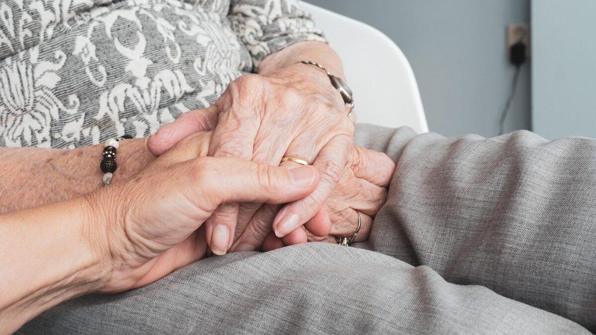Gros plan sur des mains âgées tenues par une autre personne, posées sur un tissu gris, avec une bague, un bracelet perlé et une manche imprimée visibles.