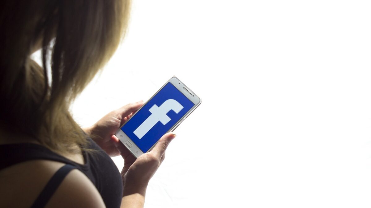 Une femme blonde, vue de dos, tient un smartphone affichant le logo bleu « f » de Facebook sur fond blanc lumineux.