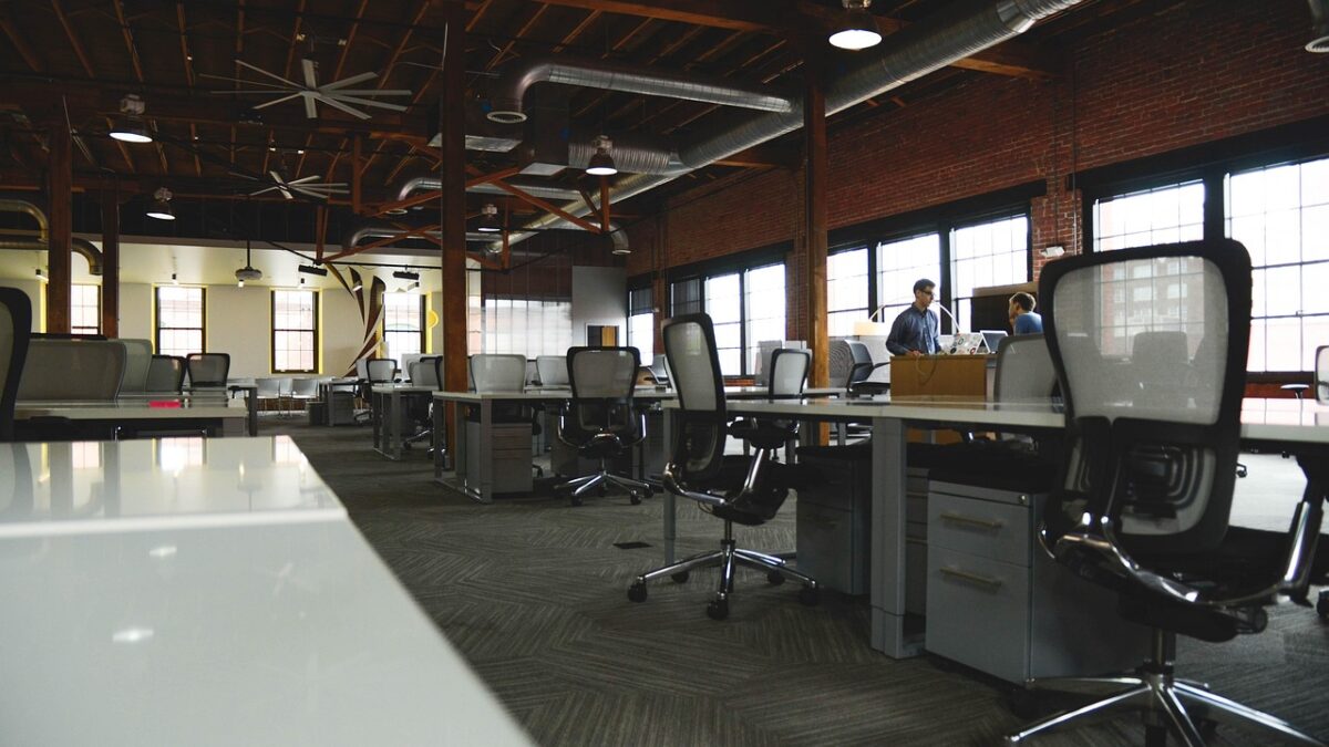 Grand open space de bureaux avec de nombreux postes de travail, chaises en maille noires, conduits apparents et murs en brique. Deux personnes sont visibles au fond, près d'un bureau.