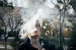 Quelles perspectives pour le marché des cigarettes électroniques face aux nouvelles régulations ? Portrait of a young man vaping in a park, releasing clouds of vapor on a clear day.