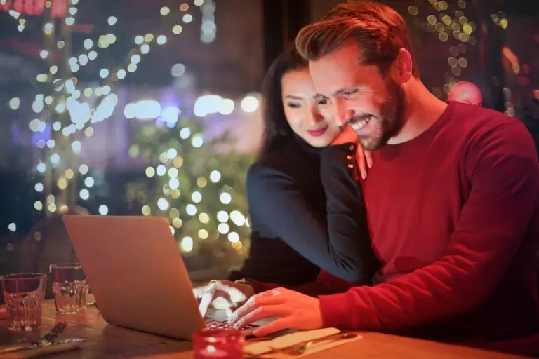 Tout ce que vous devez savoir sur le chat vidéo Bazoocam A couple using a laptop at a cozy restaurant with festive bokeh lights, enjoying online shopping together.