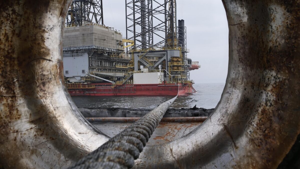 Plateforme pétrolière offshore en mer avec un navire rouge à proximité, vue à travers un anneau d'amarrage rouillé au premier plan, relié par un cordage.