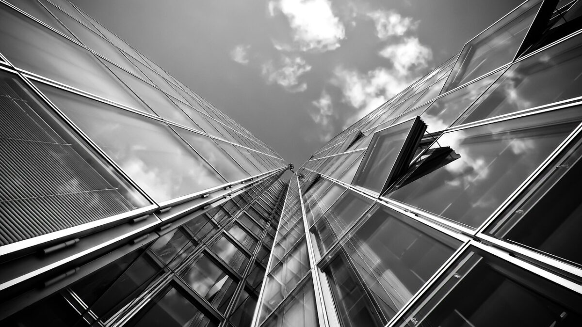 Tour en contre-plongée aux façades de verre et montants métalliques, se détachant sur un ciel nuageux, en noir et blanc.
