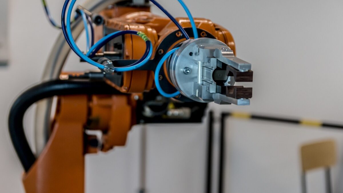 Bras robotique industriel orange à six axes, avec pince métallique grise et câbles bleus, devant un mur blanc ; une chaise et une barrière noire et jaune sont visibles à l'arrière-plan.
