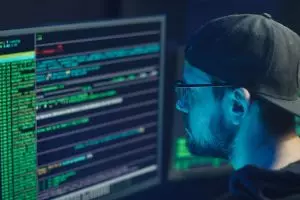 Comment reconnaître et éviter les arnaques en ligne en 2025 ? A cybersecurity expert inspecting lines of code on multiple monitors in a dimly lit office.