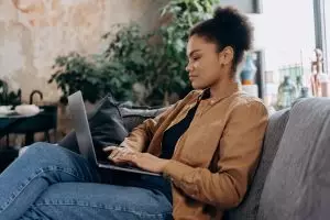A young woman in casual clothing works on her laptop in a cozy home office setting.