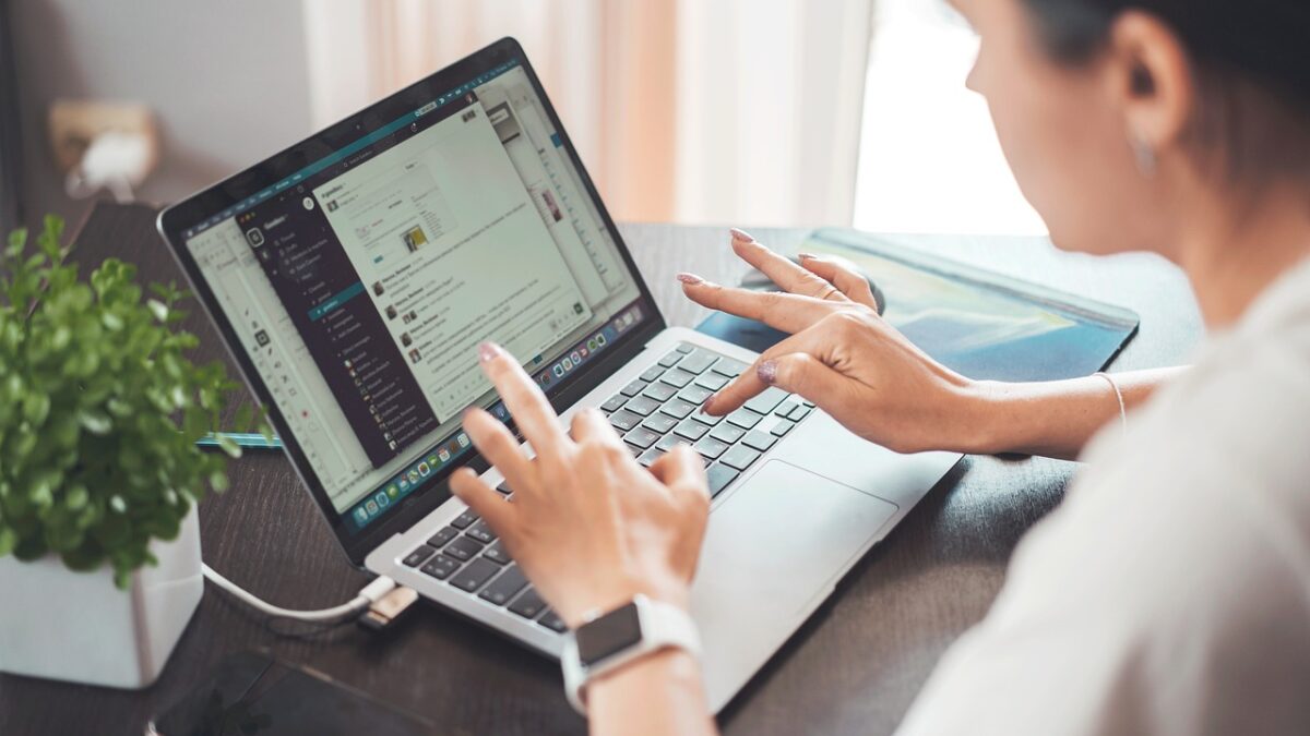 Une femme tape sur un ordinateur portable posé sur un bureau, à côté d'une plante verte et d'un téléphone. L'écran affiche une interface de messagerie.
