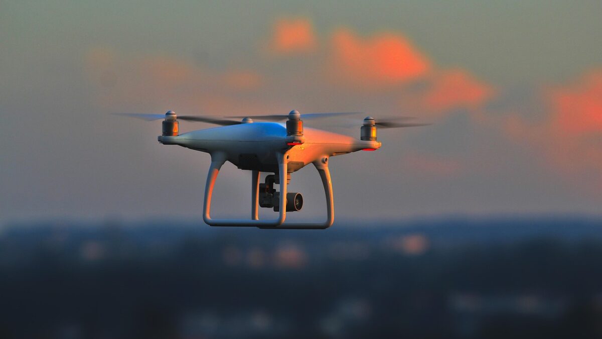Un drone quadrirotor blanc en vol au coucher du soleil, caméra fixée sous le fuselage, sur fond de ciel orangé et de paysage urbain flou.