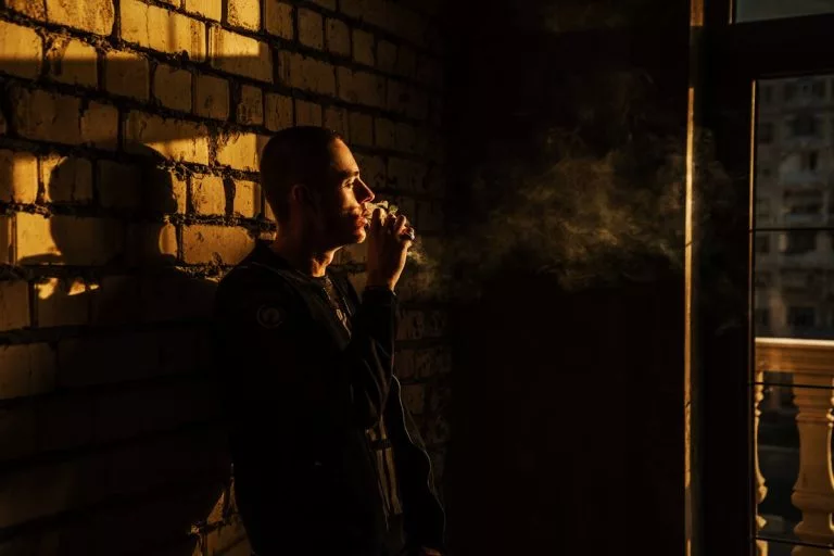A thoughtful man smokes in a dimly lit urban setting, casting shadows on a brick wall.