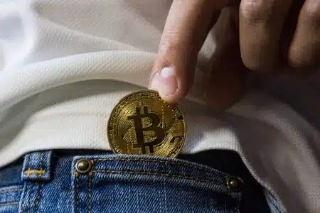 Comment l’adoption crypto ouvre de nouvelles portes d’accès aux loisirs Close-up of hand putting Bitcoin in jeans pocket symbolizing cryptocurrency savings.