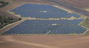 Vue aérienne d'une grande centrale solaire avec des rangées de panneaux photovoltaïques bleus sur un terrain agricole, quelques petits bâtiments techniques et une route en bordure.