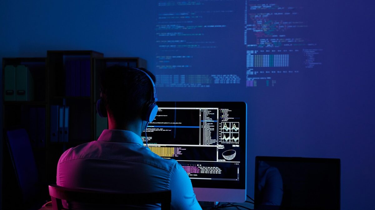 Un homme de dos, casque audio sur la tête, travaille dans une pièce sombre éclairée en bleu et violet devant un grand écran affichant du code et des graphiques, avec un ordinateur portable à sa droite.