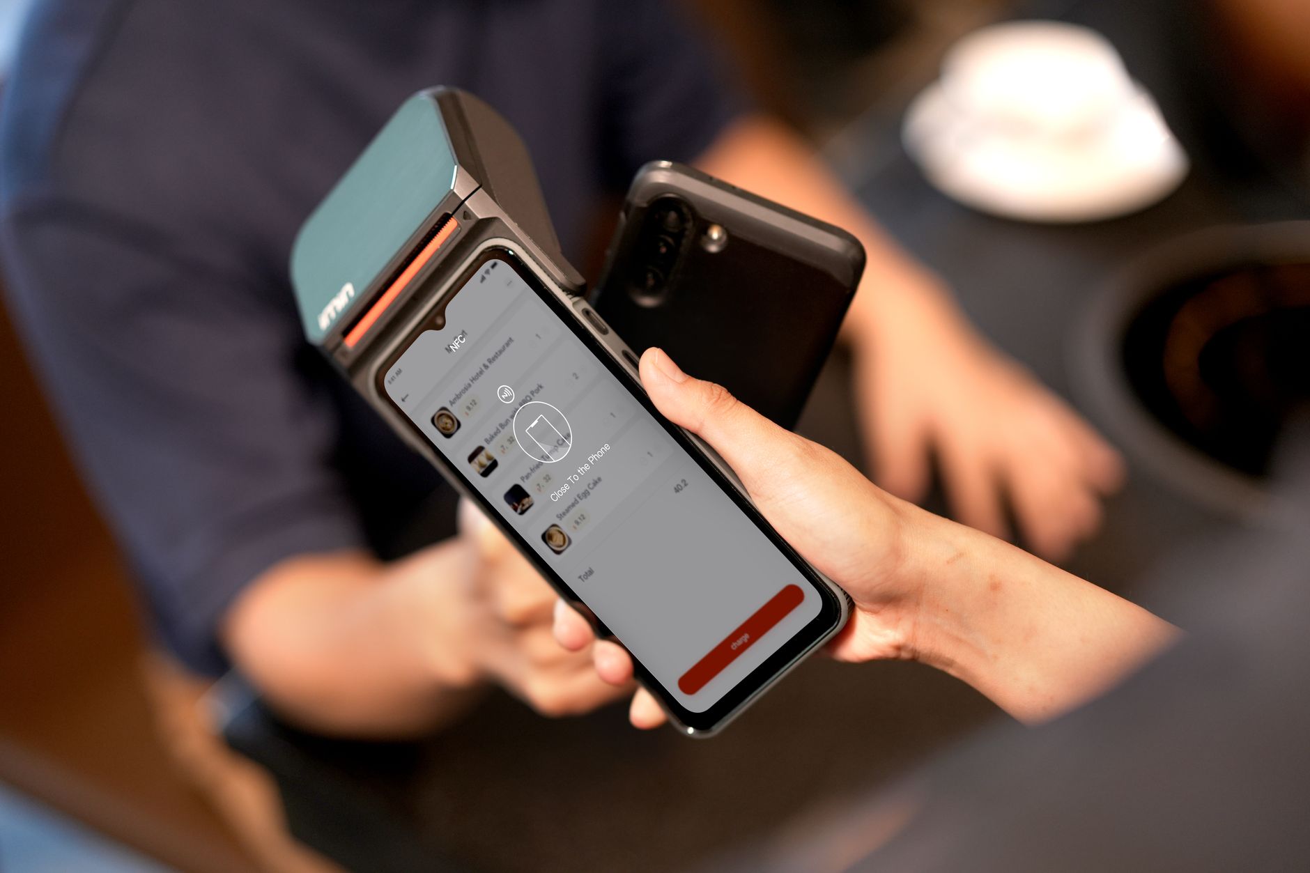 Une main approche un smartphone d’un terminal de paiement portable dont l’écran affiche une commande, dans un café flou.