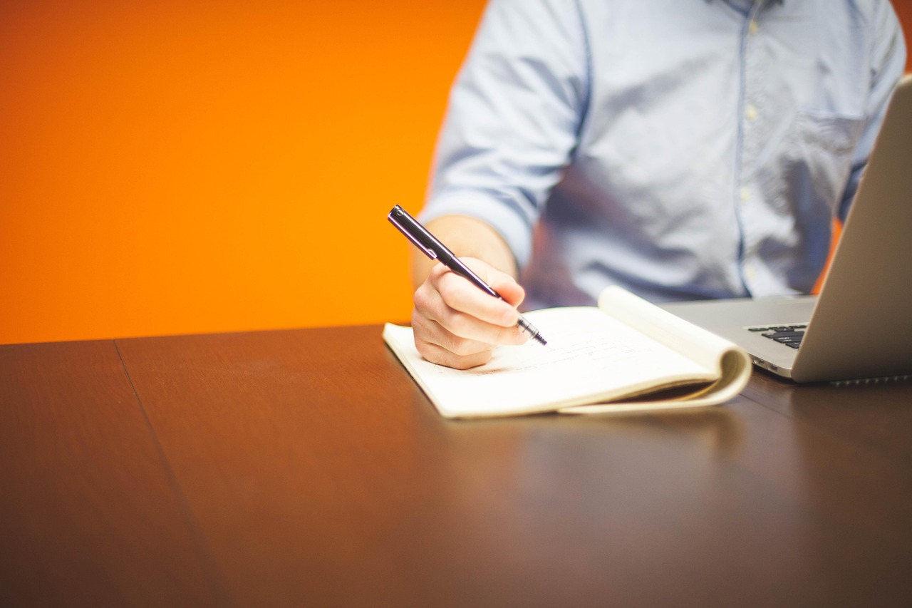Une personne prend des notes à la main dans un carnet à côté d'un ordinateur portable, sur une table en bois.