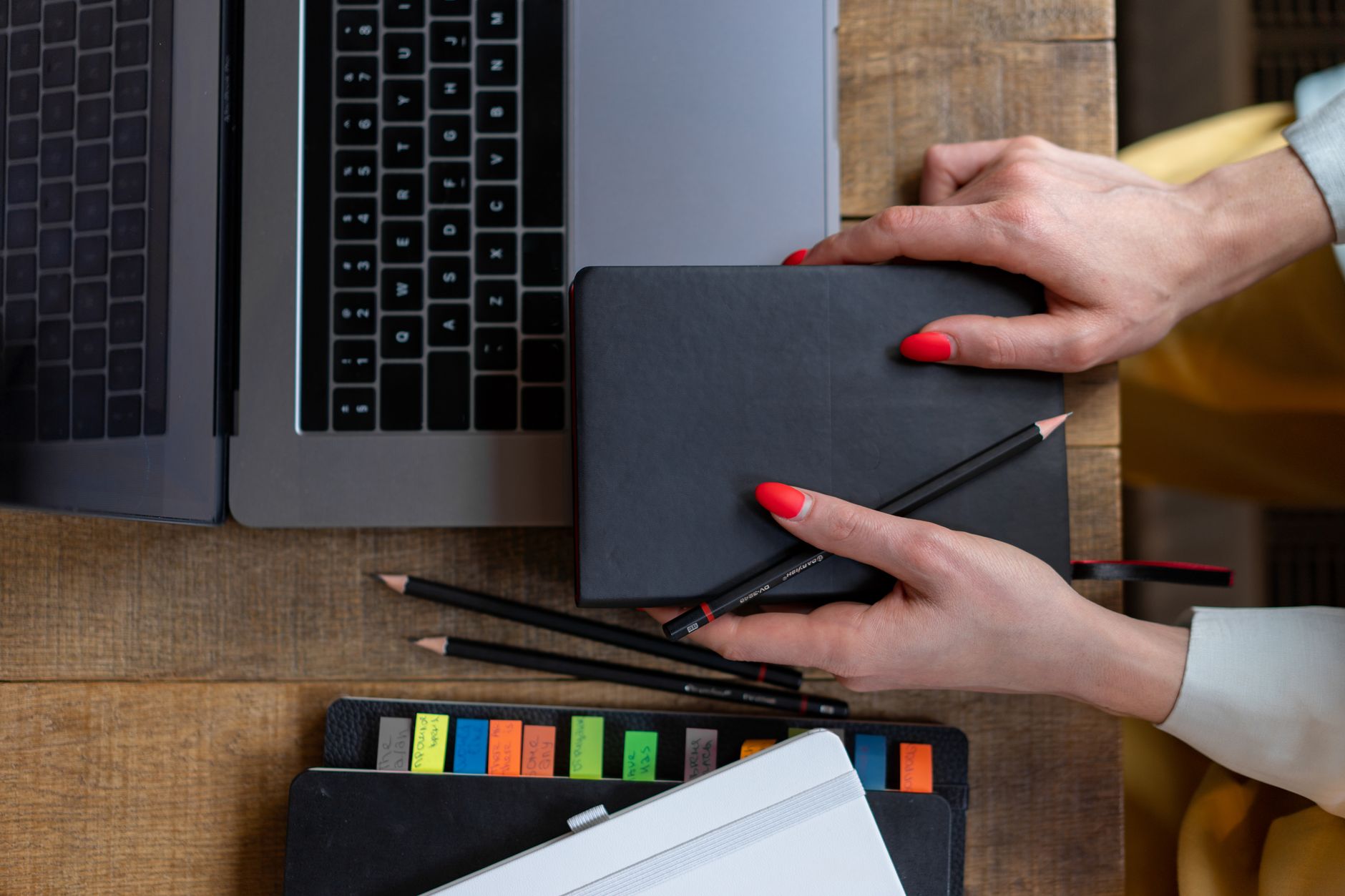Vue de dessus d’un bureau en bois avec deux ordinateurs portables, des mains tenant un carnet noir, des crayons et des carnets.